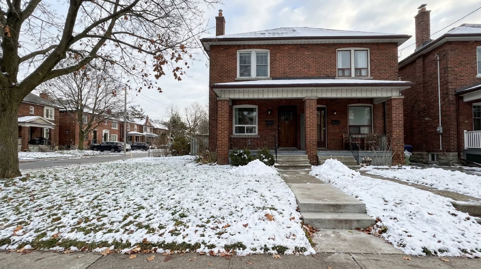 A red brick Toronto semi-detached rental house in early winter, with a small front lawn under a light dusting of snow and a freshly shovelled front walk and steps