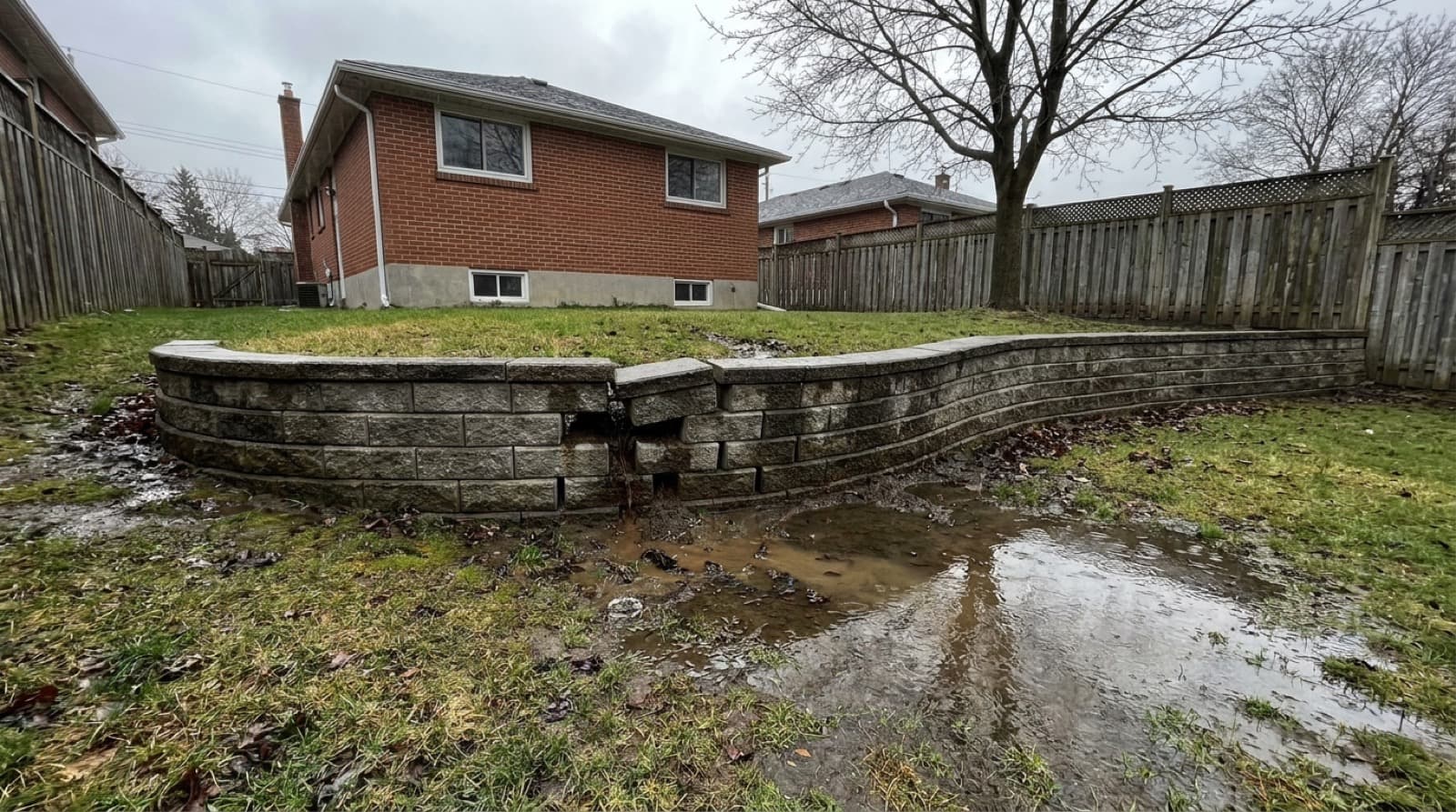 A segmental concrete block retaining wall leaning and bulging outward in a sloped Toronto backyard after heavy rain, under an overcast sky